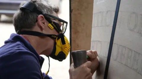 A person wearing a heavy-duty mask chisels out letters drawn out on a sheet.