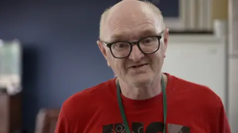 A man with glasses is looking at the camera. He is wearing a red tee shirt with a logo on the front.