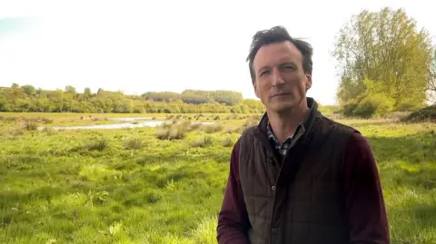 Tim Field is wearing a brown gilet over a purple sweater and checked shirt. Behind him a field is overgrown and a river winds through the middle.