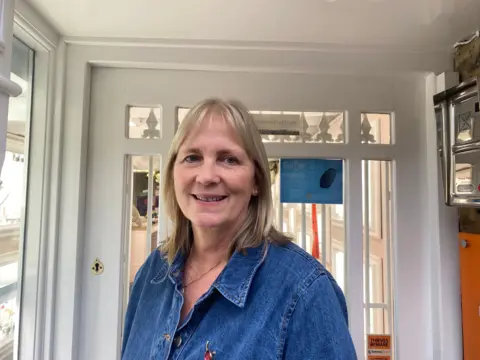 Heidi stands at the entrance to her shop, wearing a blue top and a red poppy pin