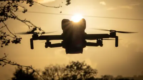 Getty Images A small drone with four propellers hovers near some trees. It is either near dawn or dusk as it appears in silhouette with the sun behind it.