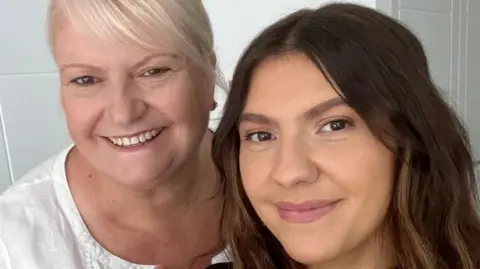 Two women pictured together. Mainly showing their heads. The woman on the left has blonde hair and is wearing a white blouse. The one on the right has dark brown hair and only her face can be seen. Both are smiling. 