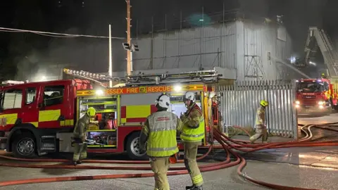 Essex County Fire and Rescue Service Two fire engines, which are red and yellow, are next to a silver warehouse. One is at the front of the building, on the left, which has four firemen around it. The other is on the side of the building, on the right, which has its aerial ladder up.