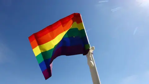 A pride flag waves in the wind. Behind it the sky is clear blue and sunlight can be seen from the top right corner of the image. The flag is striped red, orange, yellow, green, blue and purple and tied to a white flagpole.