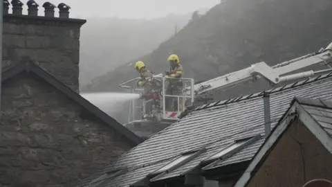 Firefighters spray water onto a building while perched on an aerial ladder with the mountainside of the area seen in the mist behind them.