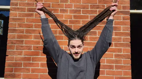A man in a grey jumper stood in front of a brick wall. He is lifting up his very long dreadlocks with both hands.