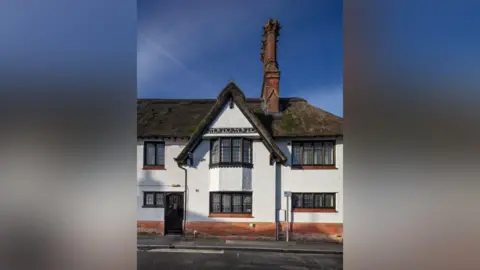Historic England Archive Part of a large white thatched building is in the middle. There is a pavement outside of the building. The windows have red brick ledges, and there is a large chimney on the right hand side of the building.
