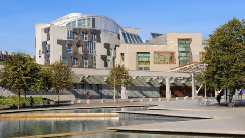 Getty Images A general view of the Scottish Parliament on a sunny day, with a manmade pond in the foreground. The stone building has wood panelling on the outside. 