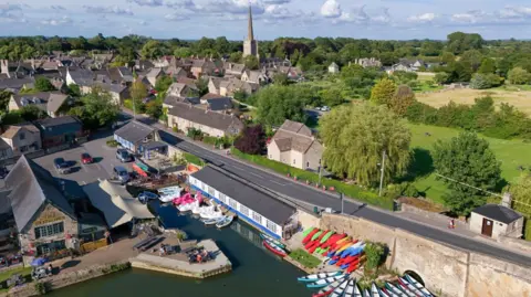 Cotswold Canoe Hire A birds eye view of a canoe hire next to the Thames. There is a long stretch of road next to it, surrounded by buildings , a car park and some greenery. 