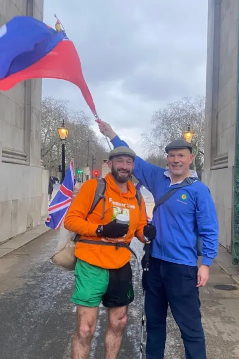 Sam Stables A man in an orange top, green and black shorts next a man in a blue top and dark trousers. The man with the blue top is waving a flag of blue and red over the other man. 