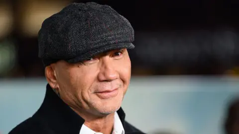 Dave Bautista smiles for cameras as he strolls the red carpet at a premiere event. He's wearing a flat cap along with a high-collared jacket over a white shirt.