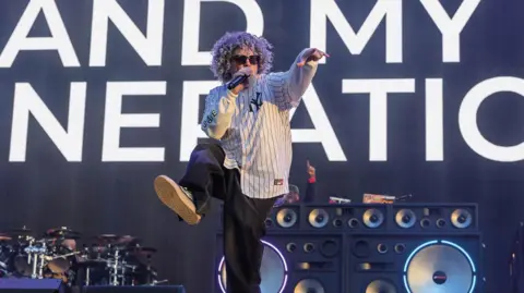 Getty Images A man wearing a white shirt and black trousers, singing into a microphone. He is pointing to the right with his left hand and kicking his right leg into the air.