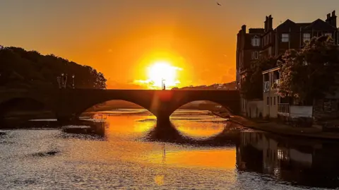 Sunset over a bridge over a river, the sun is reflected on the water.