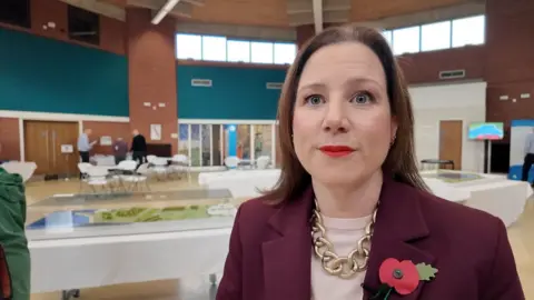 Ms Dubois has dark hair, wears a burgandy jacket and gold chain and stands in front of a model of the reservoir.
