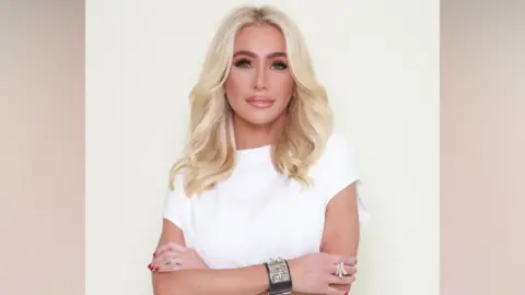 Studio portrait of a woman with long blonde hair, wearing a white top and jewellery, standing against a plain light background with arms crossed. She wears a black band around her wrist