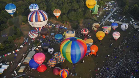 PA Drone shot of dozens of balloons lifting off the ground in a field as a large crowd watches. The balloons are of all different shapes and colours, including bulldogs, a rocket ship, and a dragon. 