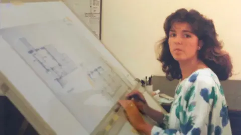 Lianne Morgan A woman with long brown hair sat in front of a board with planning building designs on it. 
