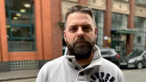 A man with brown hair and a beard is wearing a grey top. He is standing in a street with a blurred red brick building and cars behind him.