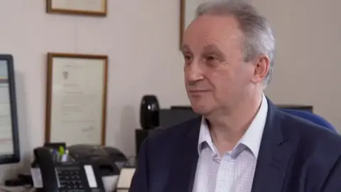 Julian Spinks is sitting in his GP surgery with his desk behind him and a computer. He has grey hair and is wearing a white and pink checked shirt and a navy blazer