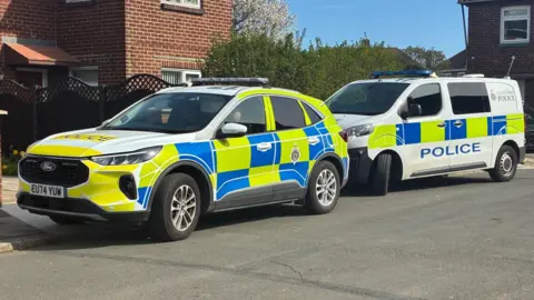 A police car parked in front of a police van. The car is a large, white Ford with bright yellow and blue squares covering most of the car. The van is mostly white with the same pattern. They are parked half on the curb in front of a red brick semi-detached house with a brown fence.