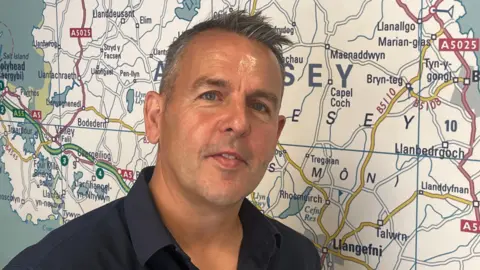Mike Williams is photographed standing in front of a map of Anglesey. He has short grey hair and is wearing a black shirt. 