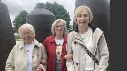 Shows three women, all with fair hair, standing in front of a piece of public art consisting of large vertical 