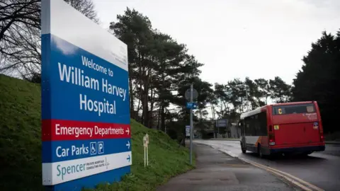 A bus drives past a blue sign. The sign reads "Welcome to William Harvey Hospital" and is on a grassy hill.