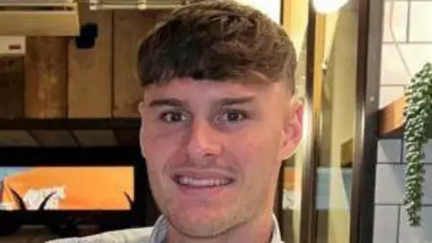 A slightly pixelated image of a young man with dark hair. He is sitting smiling in a restaurant and wearing a dress shirt. 
