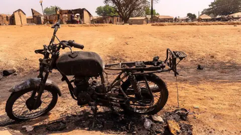 The charred frame of a burnt motorbike sits on dry, yellow grass near a small rural village.