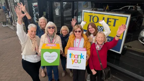 Seven women are standing outside a black shop with a white sign that reads "bodycare". There are large glass windows and a large yellow poster that reads "to let". The women have their hands in the air in celebration, and two of them are holding colourful boxes with the charity logo on the front.