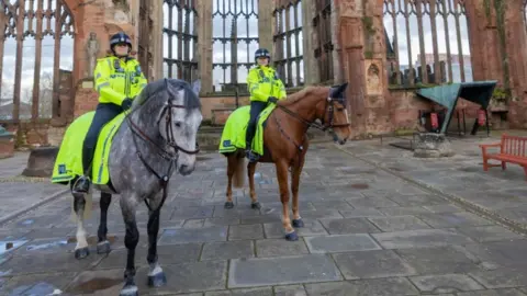 Two police officers in bright yellow reflective clothing and black riding helmets, one on a grey coloured horse and the other on a brown horse with the remains of church windows in the background