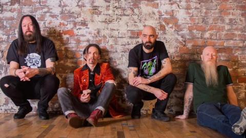The four members of the band Space pictured sitting on a wooden floor with a brick wall behind them