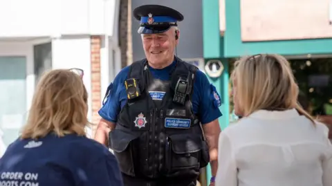 Essex Police Allan is wearing the uniform described above and chatting to three people who we can see from behind. He is smiling. They are outside a shop with turquoise decour.