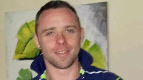 A headshot of murder victim Shane Whitla, who has short brown hair and stubble and is wearing a blue, green and white polo shirt