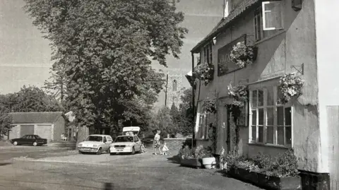 Pennoyer Museum Collection A black and white photograph of the pub with hanging baskets, a woman and a child outside. Two 1980s cars are parked outside. One has its boot open.