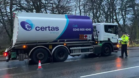 A police officer stands near the cab of a fuel tanker. It is raining and the road is wet.