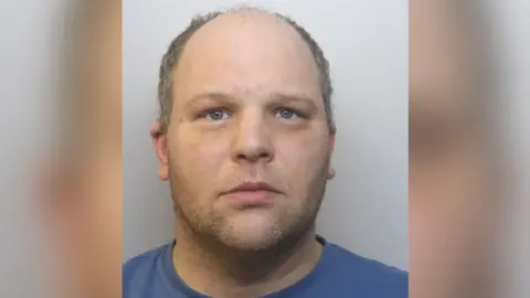 Avon and Somerset Police A balding man with stubble and greyish hair is seen from his shoulders up. He is wearing a blue T-shirt.