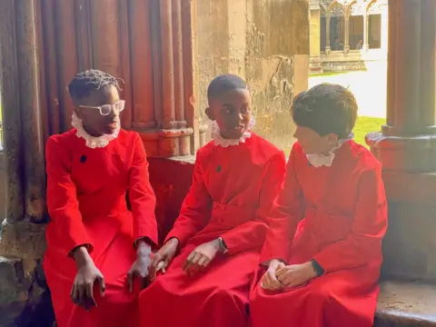Three choristers dressed in red robes chatting on a windowseat.