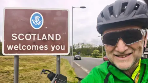 A picture of Graham Beck wearing a green jacket. He is standing in front of a brown sign that says: Scotland welcomes you. He is wearing black glasses and a helmet.