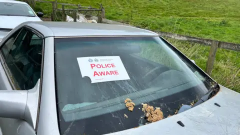 A silver car parked near a grassy area with a wooden fence, displaying a “Police Aware” notice from Bailiwick of Guernsey Law Enforcement on its front windshield