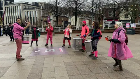 A group of people dressed in pink clothing are playing a variety of instruments on a picket line outside Sheffield Hallam University