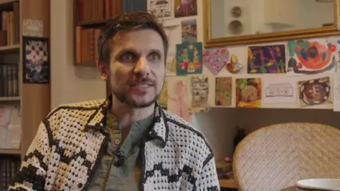 Adi Lapadatescu speaks to camera from his home. Greetings cards can be seen on the wall behind him