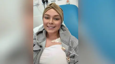 Ellie Philpot Ellie is sat on a blue hospital chair with tubes attached to her chest. She wears makeup and a brown turban covering her head. She is smiling. She wears a white t-shirt and a hoodie.