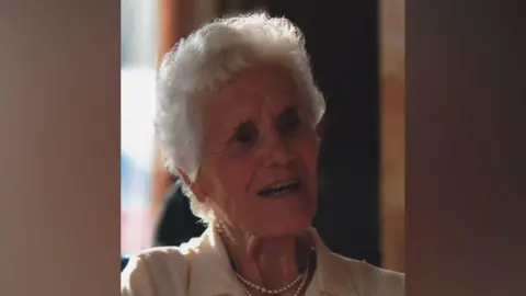 Margaret Donaldson, who has short white hair and is wearing a white pearl necklace, is smiling and looking to the side of the frame.