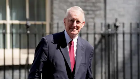 Hilary Benn outside 10 Downing Street. He has grey hair and glasses and is wearing a dark suit with a red tie.