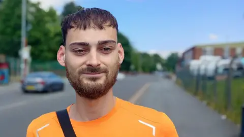 Mason Robinson, wearing an orange T-shirt and bag, speaks to the BBC in the street.