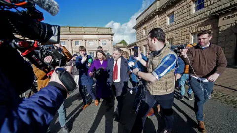 PA Media Media surround MP Mike Amesbury as he leaves court. Reporters are holding microphones towards the MP, who is wearing a dark blue jacket over a white shirt and red tie.