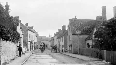 An historical photograph of a wide street in the town of Charlbury, Oxfordshire, taken around the end of the 18th Century. A man can be seen leading a horse and cart, and another leaning against a wall. 