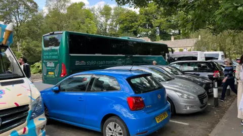 A green coach drives past a row of parked cars and an ice cream van in the Cotswolds village of Bibury. A honey coloured Cotswold stone pub can be seen in the background and there are trees everywhere.