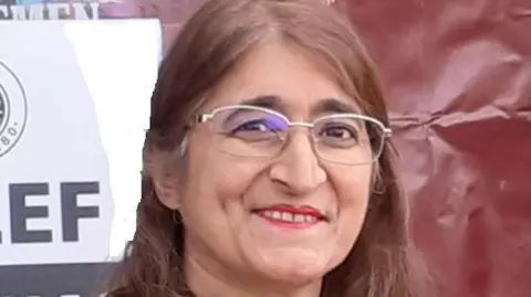 Northamptonshire Rights and Equality Council A woman smiling at the camera. She has long dark hair and is wearing glasses. She is standing in front of a banner.